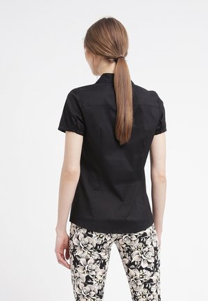 Femme aux longs cheveux bruns attachés en queue de cheval, portant un haut noir à manches courtes et un pantalon à fleurs noir et blanc, debout, de dos.