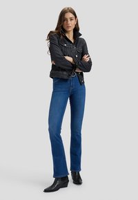 Femme aux longs cheveux bruns portant une veste en jean noire, un jean bleu et des bottines noires, debout les bras croisés sur un fond gris.