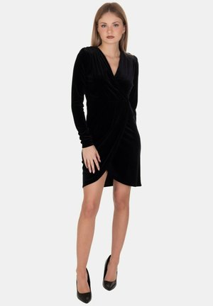 Abito avvolgente in velluto nero con maniche lunghe, scollatura a V e spacchi alta sulla coscia. Abbinato a tacchi neri a punta. Texture liscia.