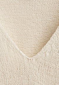 Haut en maille à col en V de couleur blanc cassé, fabriqué à partir d'un tissu léger et texturé. Présente un tissage lâche et des détails de couture subtils.