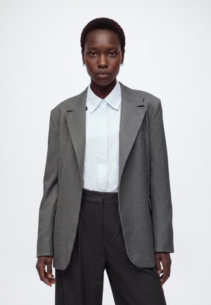 Femme aux cheveux courts portant des boucles d'oreilles créoles argentées, un blazer gris par-dessus une chemise blanche à col, et un pantalon gris foncé sur fond blanc.