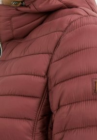 Maroon puffer jacket with a quilted texture. Features a hood and small leather tag on the side. Smooth, lightweight, and insulated materials.