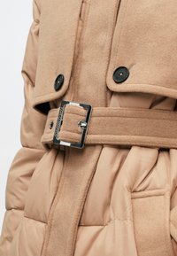 Abrigo acolchado marrón claro con una capa exterior de lana texturizada, botones negros y un hebilla plateada que acentúa la cintura.