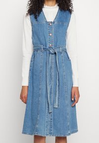 Denimmekko, jossa on tiukka yläosa, A-linjainen hame ja sivutaskut. Varustettu ruusukultaisilla napilla ja vyötäisyys. Vaalea sininen väri ja sileä rakenne.