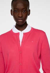 Cardigan fucsia con texture a coste, dotato di un colletto bianco e chiusura con bottoni frontali. Piccolo dettaglio del logo sul lato sinistro.