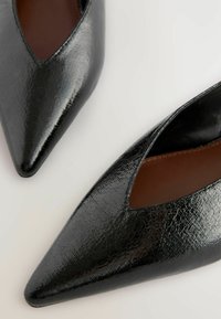 Svart lackläder pumps med spetsig tå och utskärningsdesign, med en glänsande strukturerad yta och en elegant siluett.