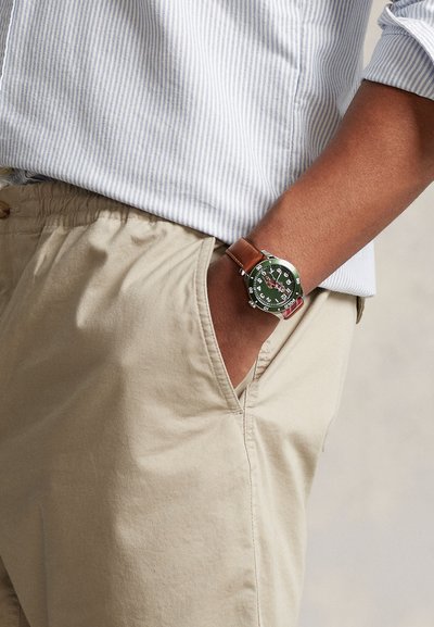 Laikrodis su žalia ciferblatu ir baltais skaičiais, nerūdijančio plieno korpusas ir ruda odinė apyrankė, nešiojamas smėlio spalvos šortų kišenėje.
