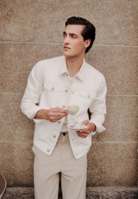 Jachetă din denim alb cu buzunare frontale, purtată peste o cămașă deschisă la culoare și pantaloni bej. Ține o cană de espresso pe un farfurioară. Fundal texturat.