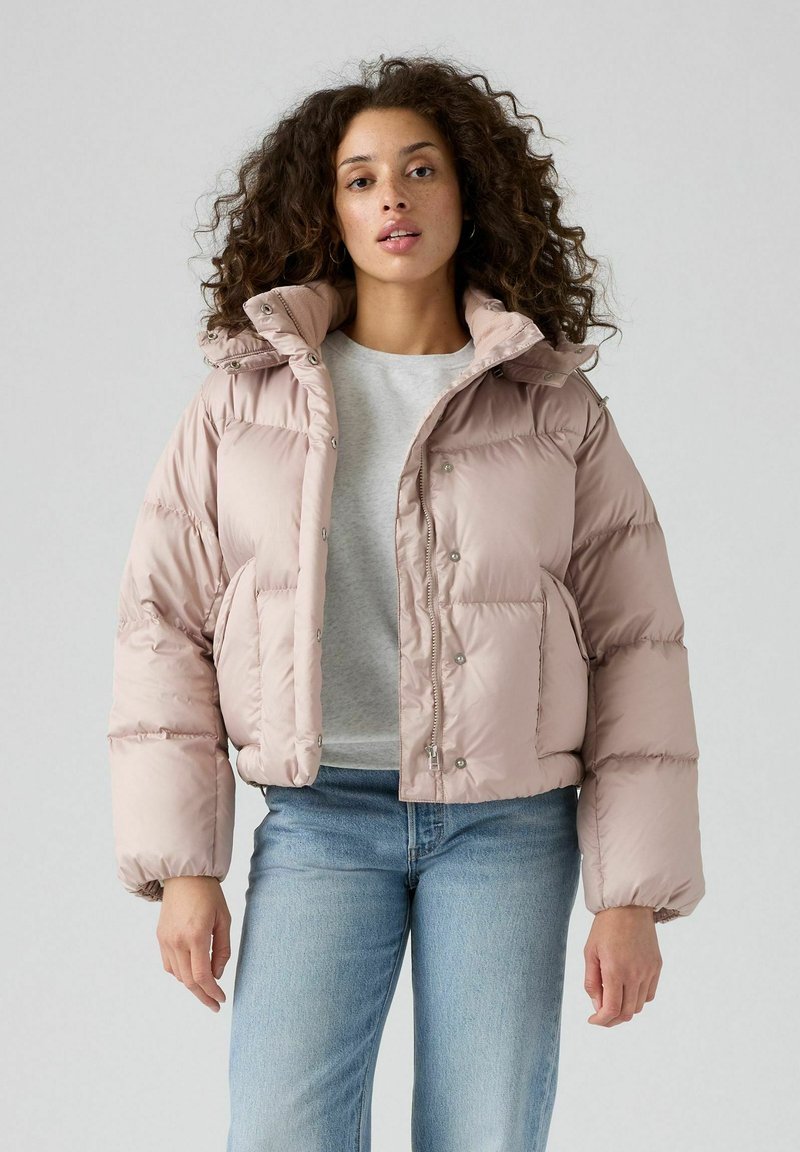 Chaqueta bomber rosa acolchada con cremallera frontal, botones a presión, bolsillos laterales y puños elásticos, combinada con una camiseta gris y vaqueros azules.