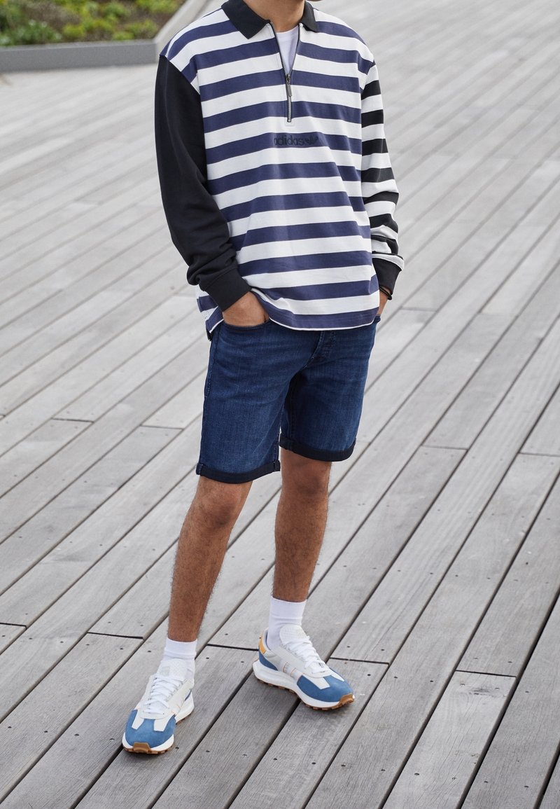 Randig tröja med dragkedja, med blå och vita horisontella ränder och svarta ärmar, kombinerad med blå denimshorts och sneakers.