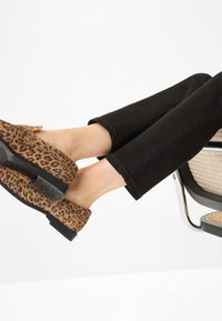 Scarpe slip-on in stampa leopardata, realizzate in suede, con suola nera, abbinate a pantaloni neri alla caviglia, posizionate su una sedia moderna.