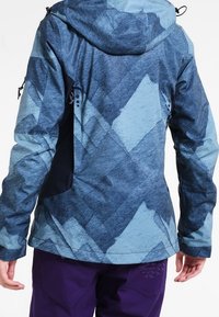 Giacca da sci blu a fantasia con un finish testurizzato, dotata di cappuccio e pannello laterale nero. Indossata con pantaloni viola, che mostrano un design moderno.
