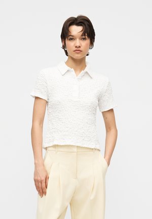 Νέος με κοντά σκουρόχρωμα μαλλιά, φορώντας λευκό μπλουζάκι με υφή, κοντά μανίκια και γιακά, σετ με κρεμ παντελόνι ψηλόμεσο, μπροστά σε γκρίζο φόντο.