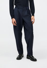 Calças de corte clássico em azul marinho, com um ajuste descontraído, apresentando pregas frontais, bolsos laterais e fecho de botão único. Usadas com sapatos pretos.