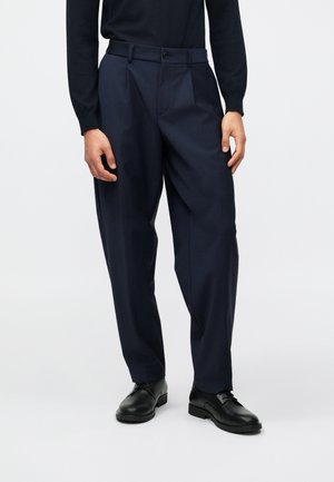 Calças de corte clássico em azul marinho, com um ajuste descontraído, apresentando pregas frontais, bolsos laterais e fecho de botão único. Usadas com sapatos pretos.