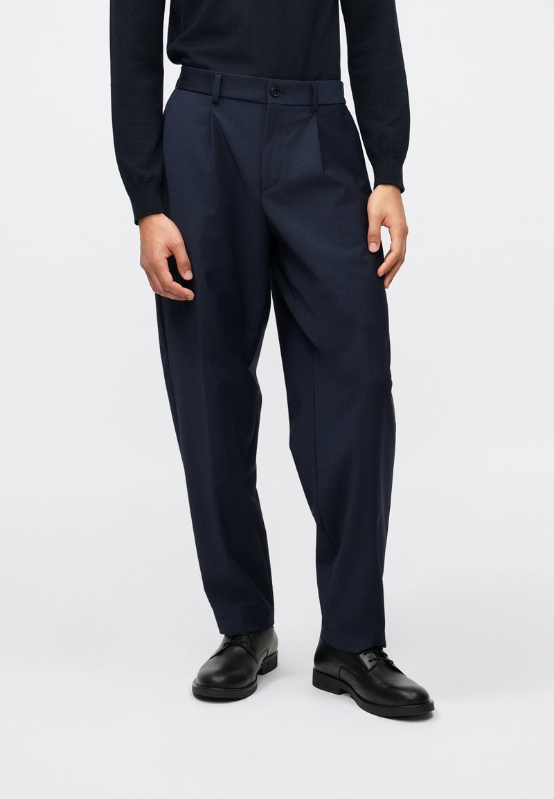 Calças de corte clássico em azul marinho, com um ajuste descontraído, apresentando pregas frontais, bolsos laterais e fecho de botão único. Usadas com sapatos pretos.