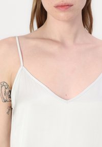 Heled hall sleeveless särk õhukeste rihmadega, V-kaelusega disain, sile tekstuur ja õrn sära. Pole nähtavaid mustreid ega riistvara detaile.
