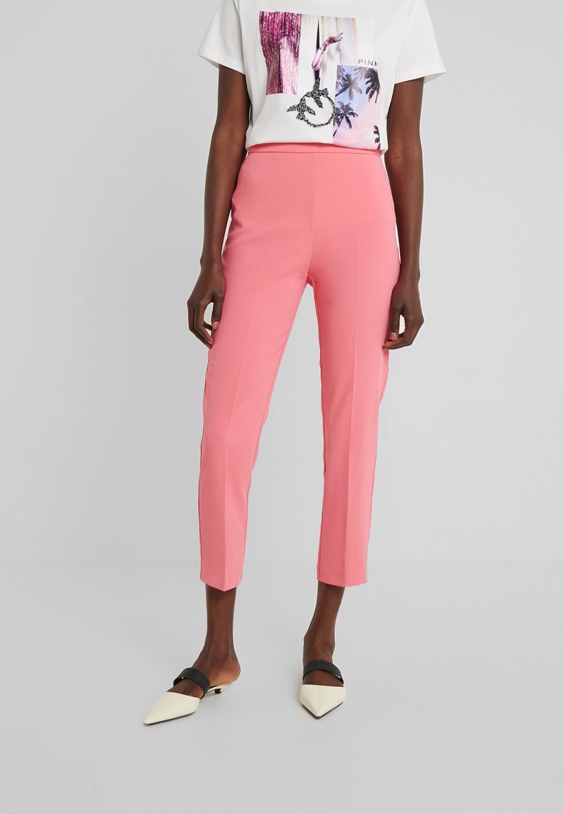 Personne portant un pantalon slim rose taille haute, un t-shirt blanc à manches courtes avec un motif graphique, et des chaussures blanches à bout pointu avec des brides noires.