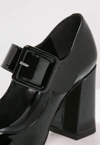 Chaussure Mary Jane en cuir verni noir avec une grande boucle carrée et un talon haut épais sur fond blanc.