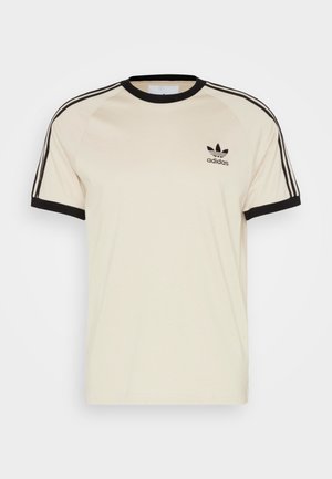 Kremowa koszulka Adidas z czarnym wykończeniem, trzema czarnymi paskami na ramionach i rękawach oraz logo Adidas na lewej piersi.