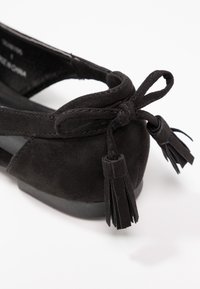 Chaussure plate en daim noir avec un nœud et des pampilles sur le talon, présentant des coutures détaillées et une semelle en caoutchouc basse.