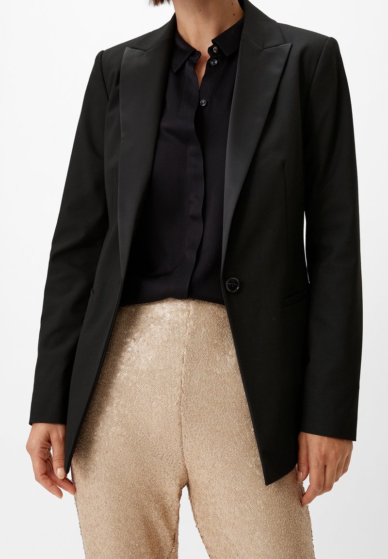 Femme portant un blazer à revers en satin noir sur une chemise noire boutonnée, associé à un pantalon beige texturé taille haute, debout devant un fond blanc.