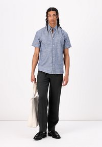Lyseblå kortermet skjorte med knapper, mørke bukser, svarte sko, holder en beige totebag. Glatt stoff med subtil tekstur.