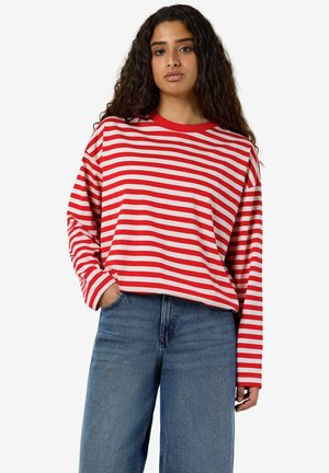 Femme aux longs cheveux bouclés portant un t-shirt à manches longues rouge et blanc rayé et un jean bleu, une main dans la poche sur fond blanc.