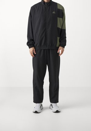 Mann trägt schwarzen Adidas-Trainingsanzug mit olivgrünen und schwarzen Blockmustern und weißen Sneakern, steht vor weißem Hintergrund.