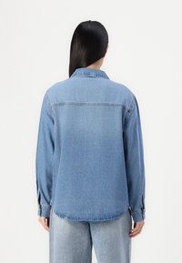 Camisa de ganga azul clara com colarinho clássico, mangas compridas e costura visível. Apresenta uma bainha ligeiramente arredondada e botões nos punhos.