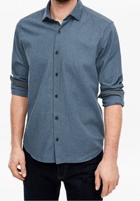 Hombre vestido con una camisa de botones azul con un patrón, mangas arremangadas y pantalones oscuros, mostrado desde el cuello hasta las caderas.