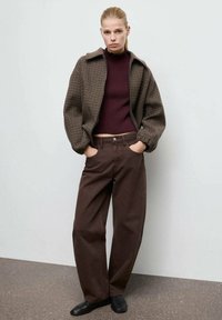 Veste en pied-de poule marron avec un col, portée par-dessus un col roulé ajusté bordeaux et un pantalon marron ample ; chaussures noires.