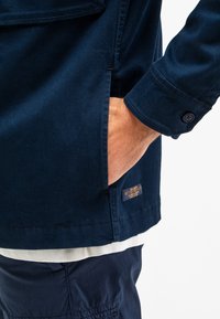 Marineblaue Jacke mit einer Seitentasche und knöpfbaren Manschetten; aus strukturiertem Stoff gefertigt; mit einem Logobranding auf dem Saum zur Markenidentifikation.