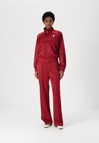 Red tracksuit made of shiny, smooth fabric. Jacket has a high collar and elastic waistband. Pants are loose-fitting with side stripes.
