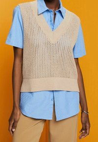 Personne portant un gilet sans manches en tricot beige sur une chemise bleu à manches courtes avec boutons et un pantalon beige, se tenant devant un fond orange.