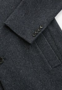Mörkgrå ullblazer med en texturerad väv, slagkragar och fyra svarta knappar på ärmen. Inkluderar en ficka på framsidan.