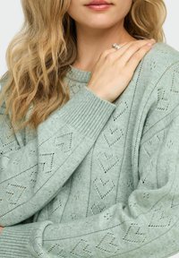 Pull en maille vert clair avec motifs de cœurs et poignets côtelés. Le modèle met en valeur le détail des épaules et une bague à la main.