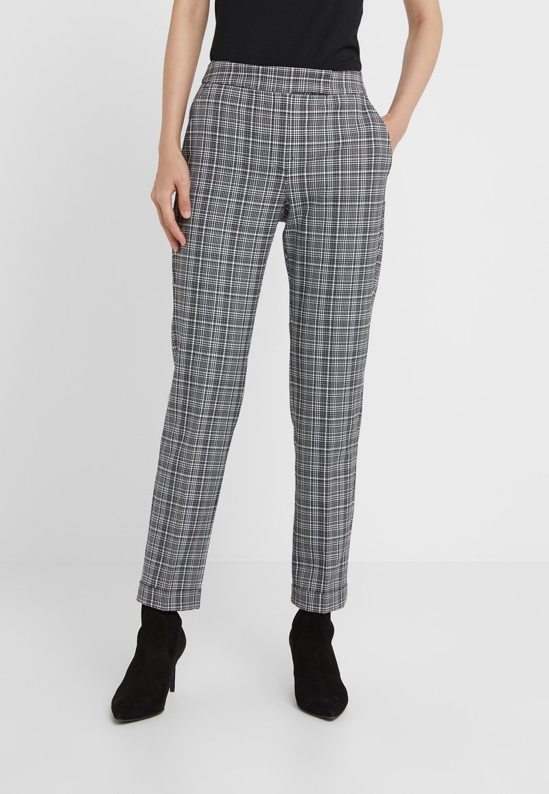 Žena v černobílých kostičkovaných zúžených kalhotách s předními kapsami a černými kotníkovými botami, stojící na jednobarevném pozadí.