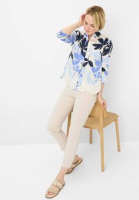 Lichtgewicht blouse met bloemenpatroon in blauw en wit; beige, slim-fit broek; tan sandalen met een gespdetail; houten stoel op de achtergrond.
