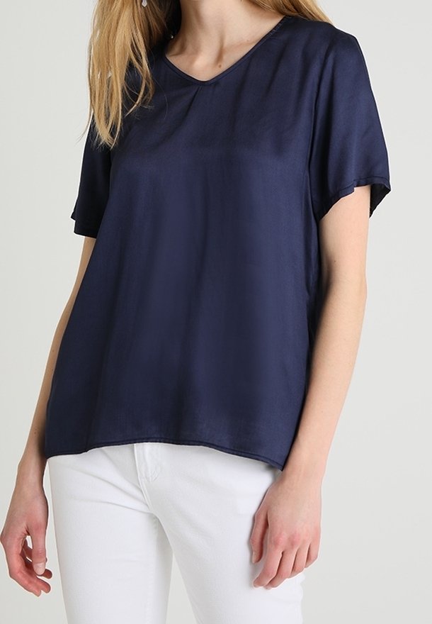 Kvinna som bär en löst sittande, kortärmad marinblå V-ringad blus tillsammans med vita byxor mot en enfärgad bakgrund.