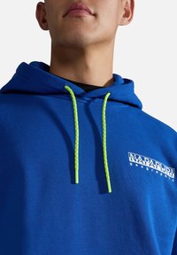 Homme portant un sweat à capuche bleu Napapijri Geographic avec des cordons vert néon visibles sur un fond clair uni.
