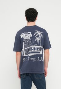 Tamsiai mėlynas medvilninis marškinėlis su balto atspaudo dizainu gale – kepsninės ženklas, palmės ir užrašas „San Diego, CA“.