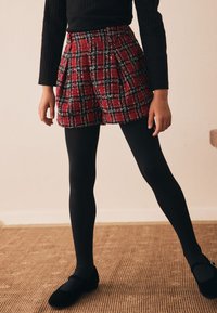 Rote karierte Shorts mit Falten, die eine Mischung aus schwarzen und weißen Akzenten aufweisen. Kombiniert mit schwarzen Strumpfhosen und flachen schwarzen Schuhen.