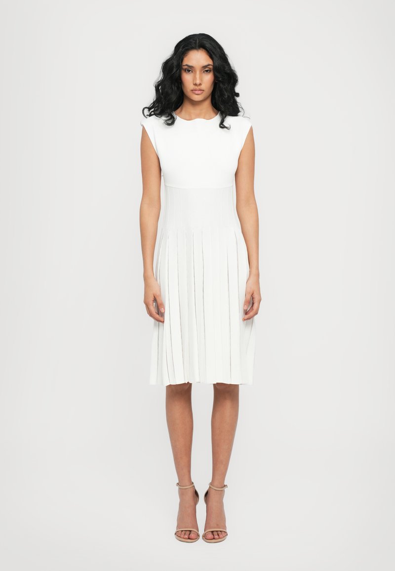 White pleated dress with a fitted bodice and cap sleeves, featuring horizontal stripes and a knee-length hemline. Worn with nude strappy heels.