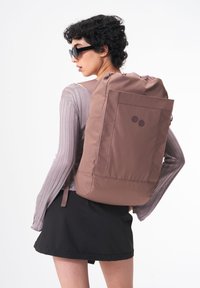 Brauner Rucksack in rechteckiger Form, aus glattem Material gefertigt, mit zwei runden Akzenten und verstellbaren Schultergurten.