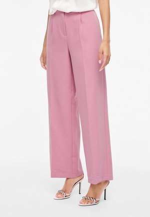 Γυναίκα που φοράει ροζ παντελόνια με ψηλή μέση και φαρδιά πόδια, σε συνδυασμό με λευκά ψηλοτάκουνα σανδάλια με ασημένια αλυσίδα.