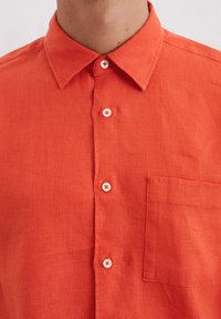 Orange linen shirt with a button-down collar, front pocket, and five white buttons featuring red accents. Smooth texture, casual design.