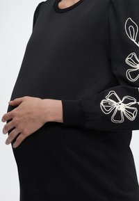 Έγκυος άτομο με μαύρο μακρυμάνικο μπλουζάκι με λευκά λουλουδάτα κεντήματα στα μανίκια, κρατώντας απαλά την κοιλιά του με το αριστερό χέρι.