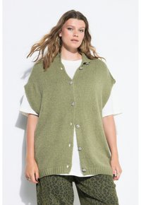 Frau trägt eine übergroße grüne Strickweste mit silbernen Knöpfen über einem weißen T-Shirt und grünen Leopardenmuster-Hosen, steht vor weißem Hintergrund.