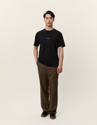 Zwarte t-shirt met korte mouwen en wit logo op de borst, gecombineerd met bruine gemusterde broek en zwarte schoenen. Neutrale achtergrond.